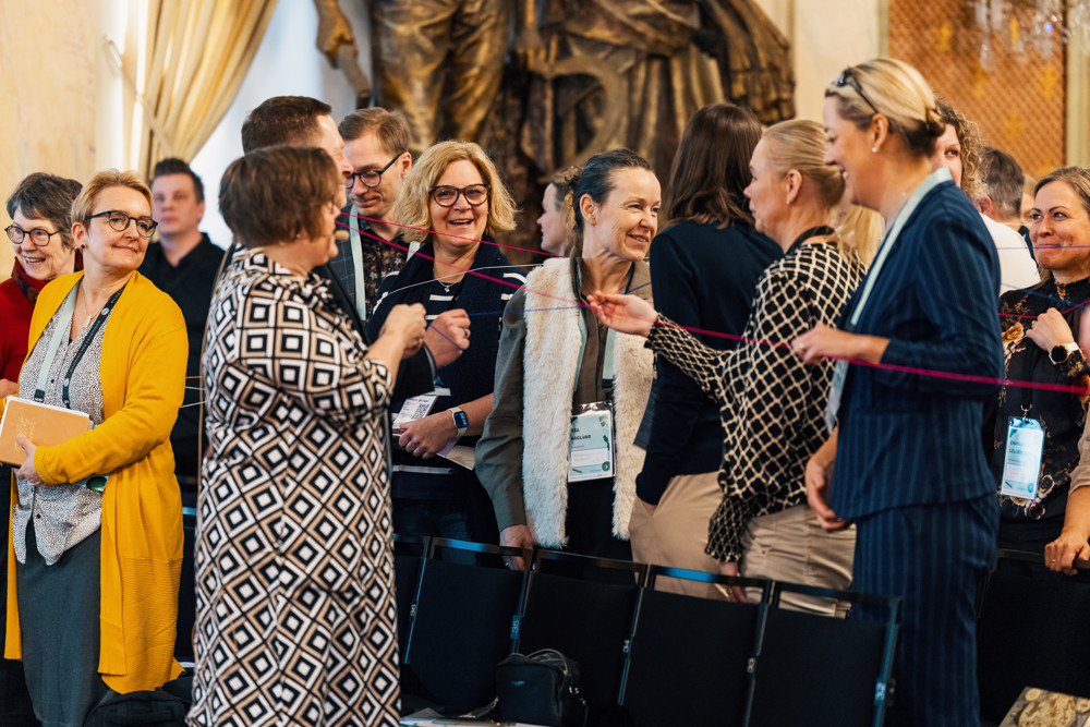På ilden syns ett tiotal människor i en konferenssal. De står upp och är vända mot varandra i olika formationer. De hae garntrådar runt sig och ser glada ut.