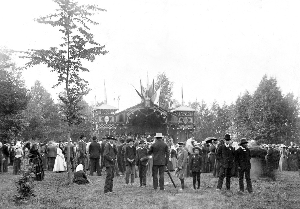 Bilden är en svartvit historisk bild från sångfesten i Ekenäs år 1891 och den hör till Sibeliusmuseumssamlingar. På bilden ses en scen i bakgrunden och en mängd människor i gammaldagskläder soms tår framför scenen som är utomhus.