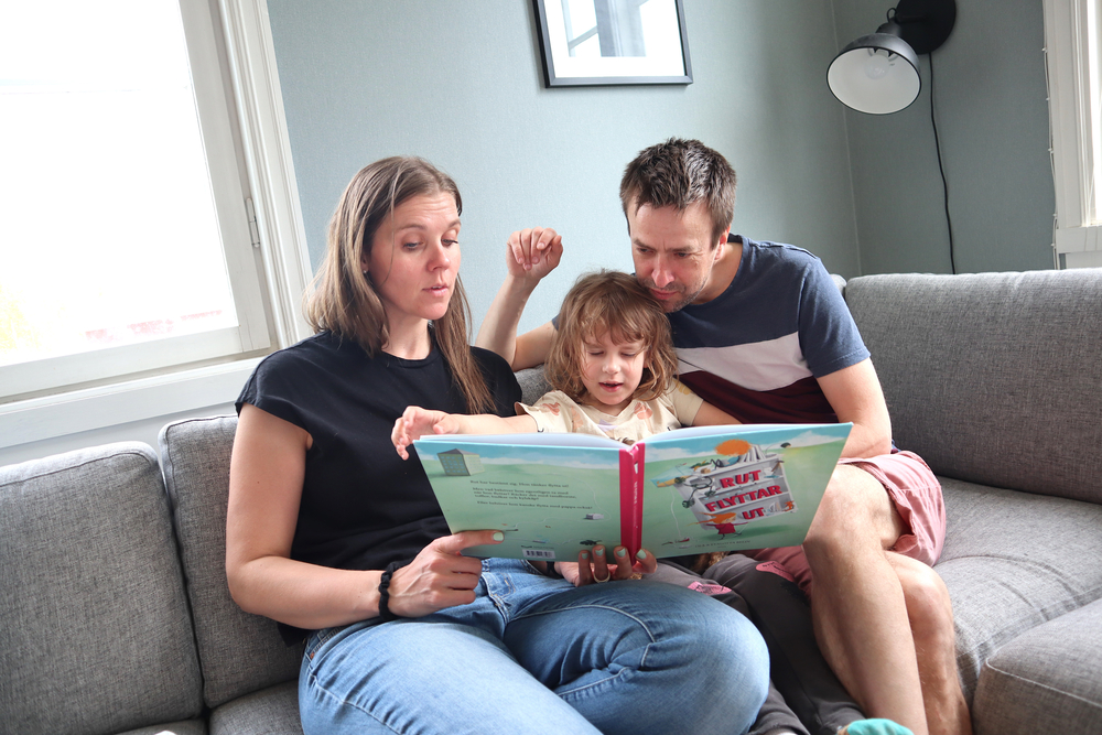 Mamma Amanda Audas-KAss och pappa Fredrik Kass läser en bok tillsammans med Hilde som sitter mellan dem i en grå soffa.