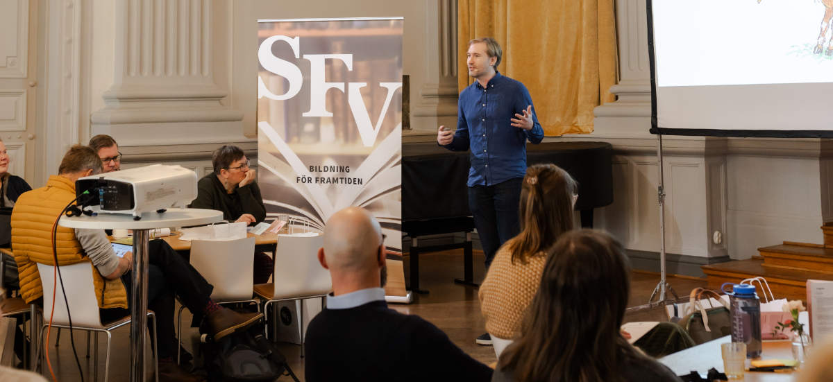 Aron Schoug talar inför publik i SFV-huset G18:s festsal. En projektor lyser upp en skärm bakom honom.