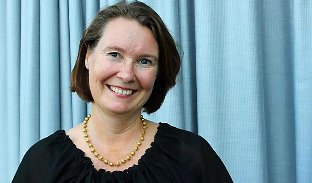 Solveig Corner ler mot kameran framför ett ljusblått draperi. Hon har ett stort guldfärgat halsband.