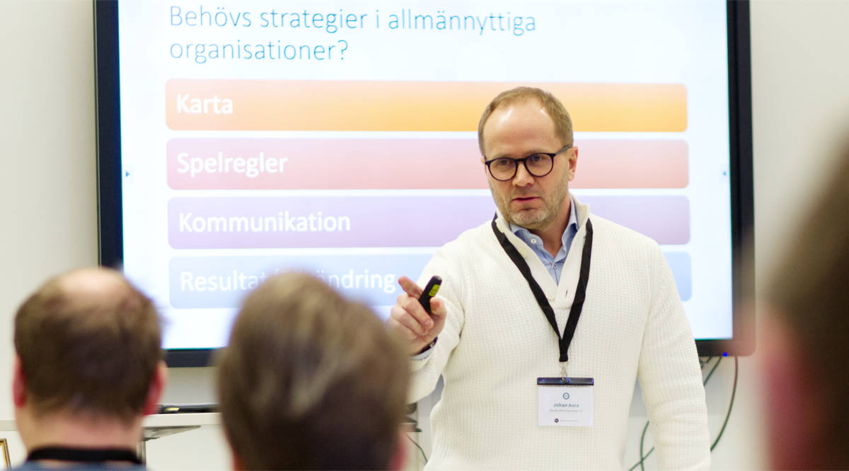 Johan Aura, klädd i en vit tröja, föreläser framför en Powerpoint om strategier i allmännyttiga organisationer. Man ser publiken bakifrån.