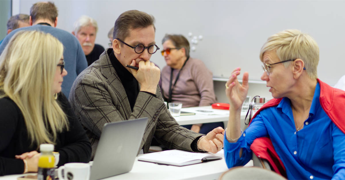 Pocke Wikström sitter vid konferensbordet och diskuterar med andra kursdeltagare. Han bär en grå kavaj över en svart skjorta.