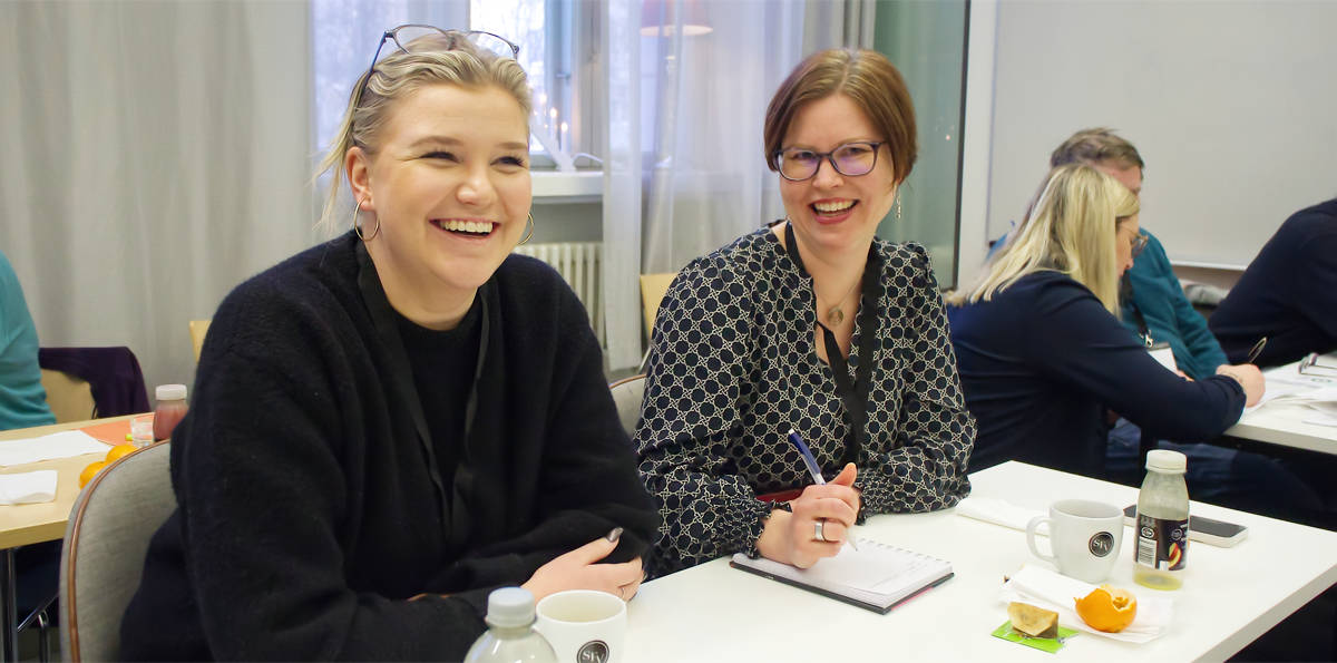 Fanny Ollus och Annika Nylander ser glada och nöjda ut. De har en kaffekopp och bulle framför sig på konferensbordet.
