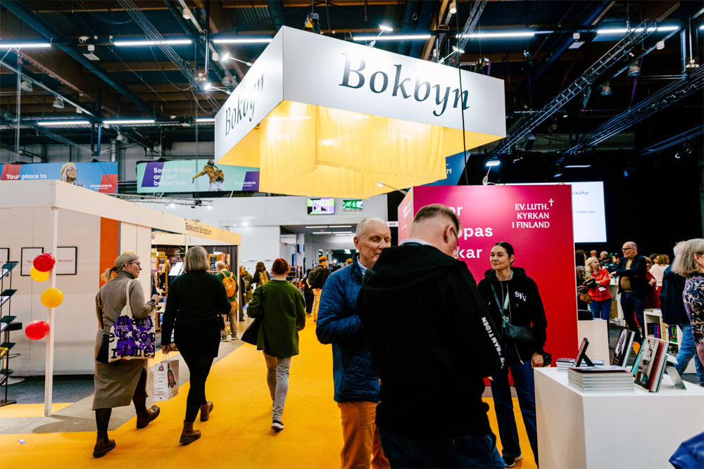 Bilden är tagen på bokmässan i Helsingfors och i taket hänger en stor skylt med ordet Bokbyn på. I förgrunden finns personer som promenerar längs en korridor och till höger två personer i svarta huvtröjor med SFV på. De diskuterar med personer framför bord med böcker.