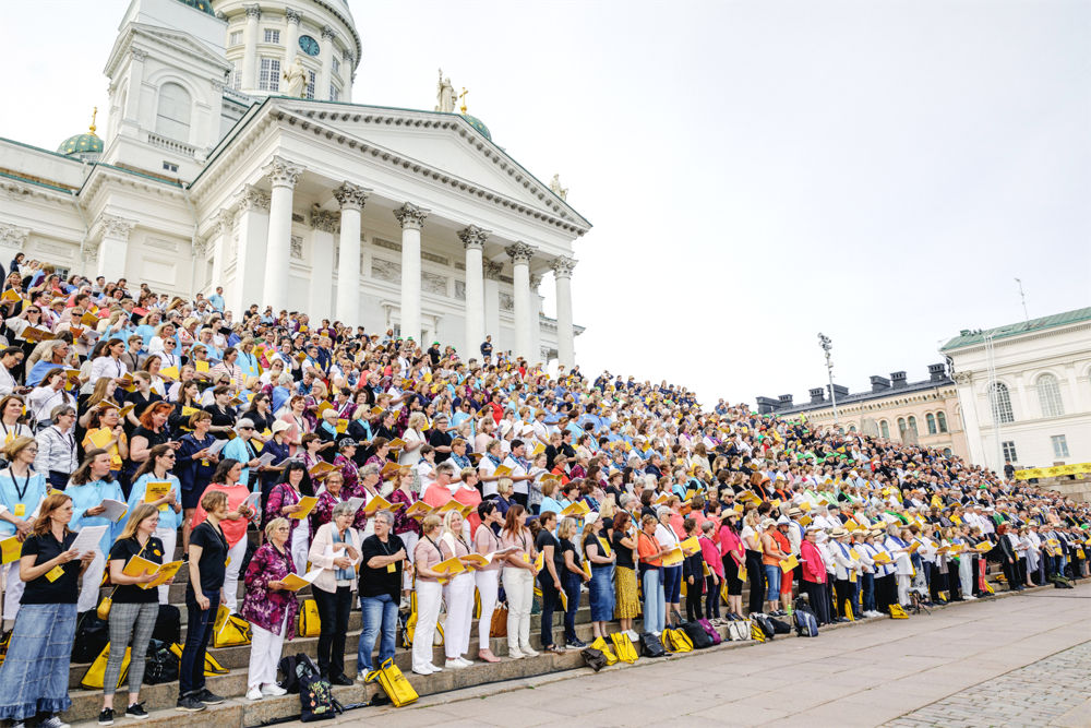 PÅ bilden ser man en massa människor soms tår uppradade på trappan till Helsingfors domkyrka. Man ser kyrkan i bakgrunden i framgrunden står människorna soma lla har knallgula påsar. De står med noter i händerna.